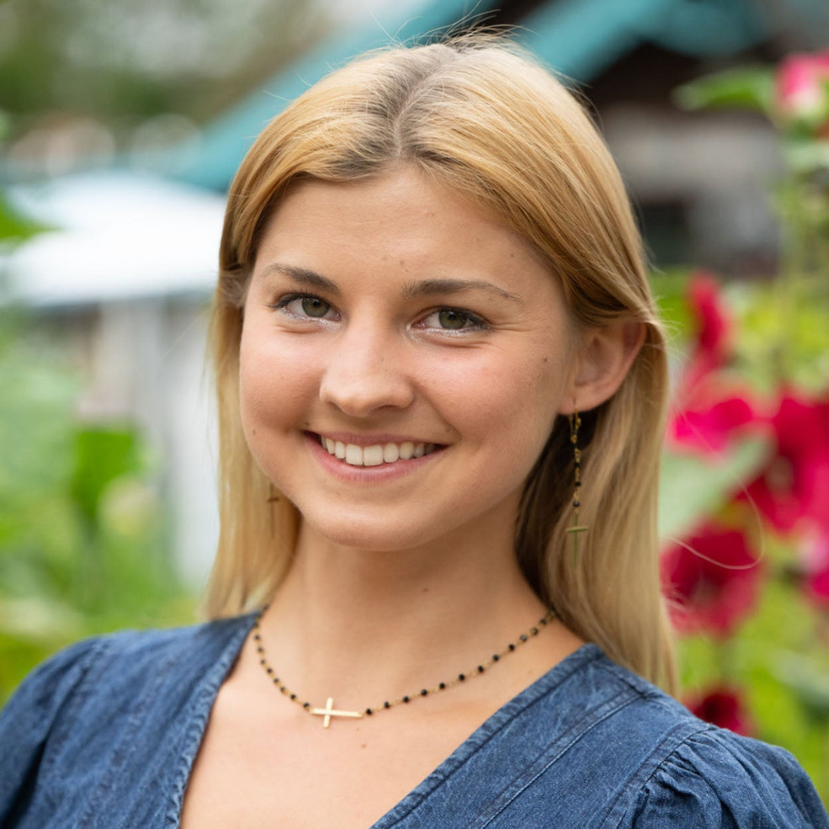 Handmade brass and black cross necklace. woman wearing a blue denim shirt with a blurred floral background